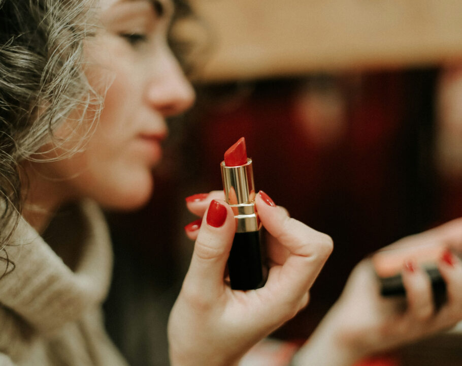 Woman with gray-ish hair and red nails holding red lipstick and looking in a makeup compact